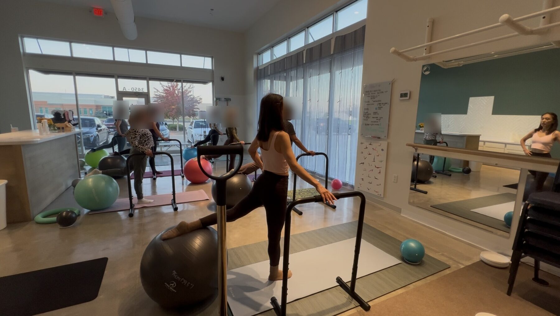 An MBB group class at IN & OUT Pilates: clients holding bars and exercise balls in lunges along the studio windows while Euna, in a pink top, leads in front of the mirror. Client faces are blurred.