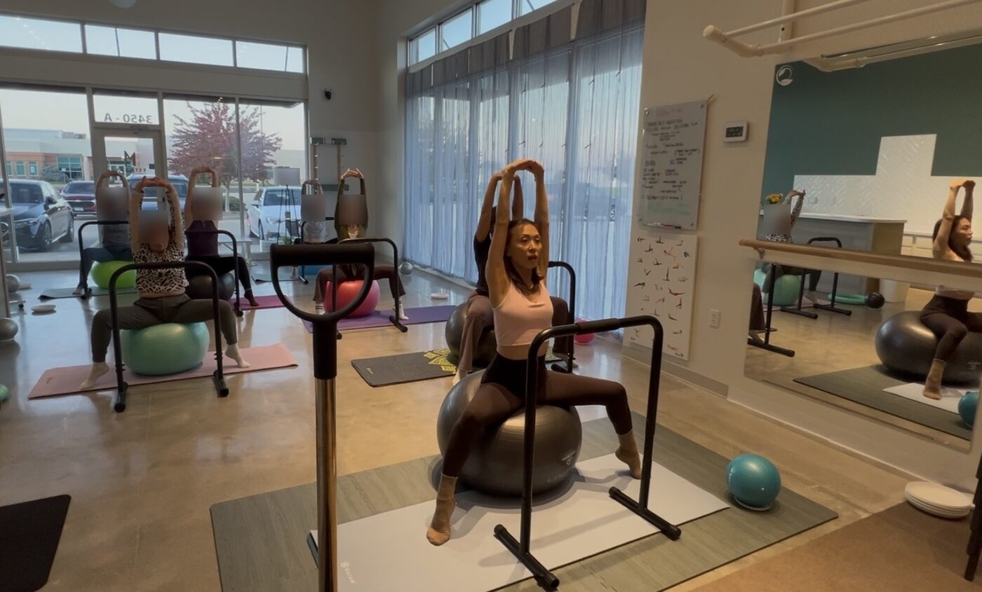 Euna leading an MBB class with arms overhead, clients seated on exercise balls behind her (client faces blurred)